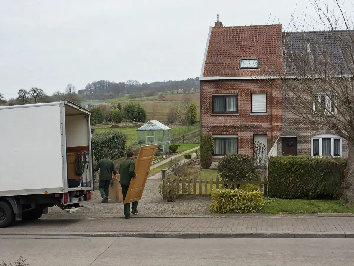 Twee mannen dragen delen van een houten kast en dozen naar een witte vrachtwagen bij een halfopen woning in Ninove